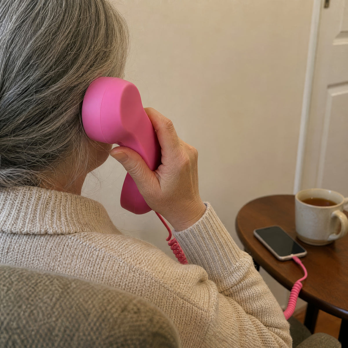 A woman in an armchair holds the pink DialBop handset to her ear, cord running to her phone on the side table